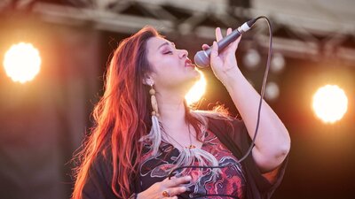 Wallabindi A female singer with long, reddish-brown hair performing live on stage under bright spotlights.