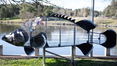 A steel wrieframe Cod sculpture with some woven textiles attached stands at the side of a lake