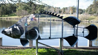 A steel wrieframe Cod sculpture with some woven textiles attached stands at the side of a lake