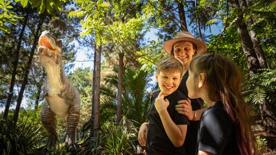 Extinction Trail - TREX A mother and two children smile in awe at the size of the animatronic T-Rex in lush fern gardens.