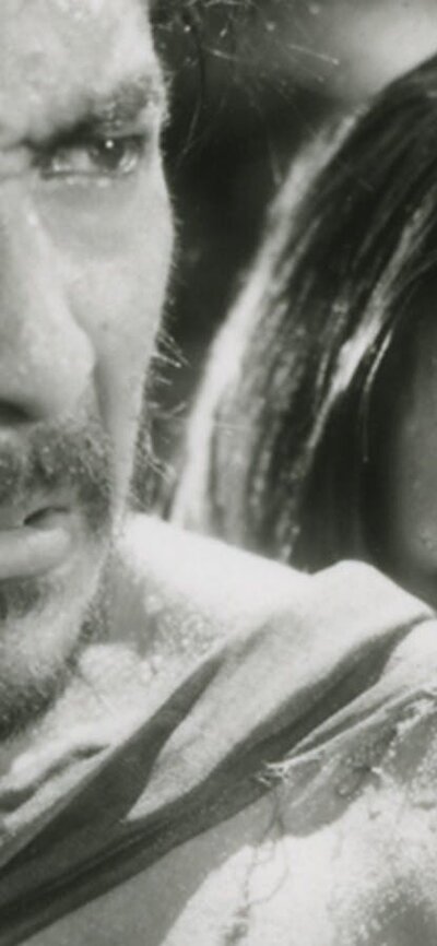 Black and white still of a man looking angry and a woman hiding behind the man