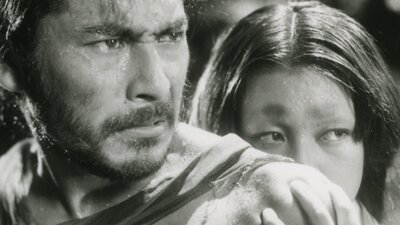 Black and white still of a man looking angry and a woman hiding behind the man