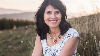 Karen Viggers A woman with dark, wavy hair sitting outdoors, wearing a white patterned top.