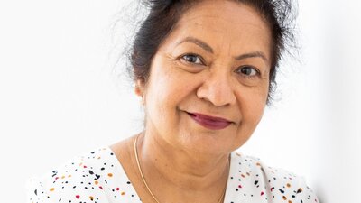 A woman smiling while resting her right hand on her left shoulder, while wearing a polka dot shirt.