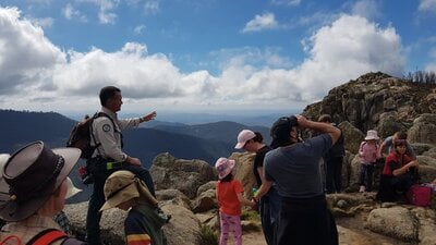 Ranger and children seated on rocks with mountain range in the background.