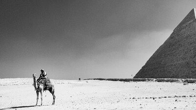 A black and white photo of a person on a camel in front of a pyramid.