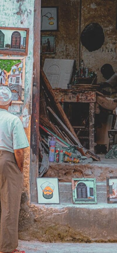 An elderly man stands in front of a small shop filled with paintings