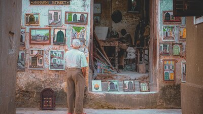 An elderly man stands in front of a small shop filled with paintings